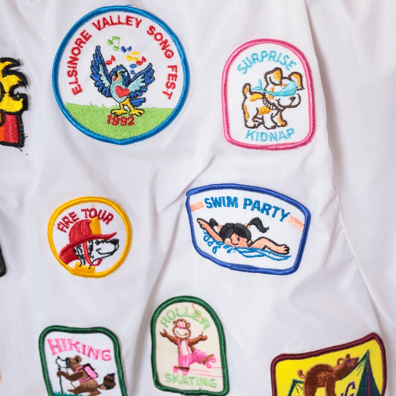 Op de afbeelding: Witte stof met kleurrijke geborduurde patches. De patches tonen verschillende thema's, waaronder een zingende vogel, een hond, een brandweerwagen, een zwembad, een rolschaatser en een wandelsituatie. De tekst op de patches omvat "Elsinore Valley Song Fest 1992", "Surprise Kidnap", "Fire Tour", "Swim Party", "Roller Skating" en "Hiking".