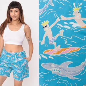 K&ouml;nnte beinhalten: Blaue und wei&szlig;e Badehose mit einem Cartoon-Druck von Surfern und Haien. Die Hose hat einen Kordelzugbund und eine lockere Passform.