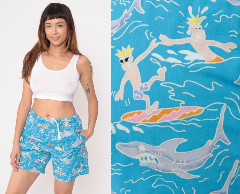 K&ouml;nnte beinhalten: Blaue und wei&szlig;e Badehose mit einem Cartoon-Druck von Surfern und Haien. Die Hose hat einen Kordelzugbund und eine lockere Passform.