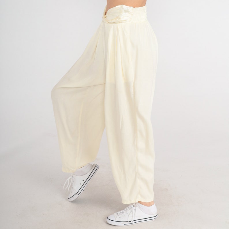May include: Cream-colored, wide-leg pants with a high, gathered waist. The pants have a relaxed fit and are paired with white sneakers. The fabric appears lightweight and flowing, suitable for casual wear.