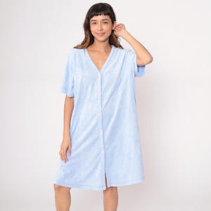 May include: A light blue terry cloth robe with short sleeves and a V-neck. The robe has a button-up front with pink buttons. The garment is knee-length and appears soft and comfortable, suitable for lounging or after-bath wear.