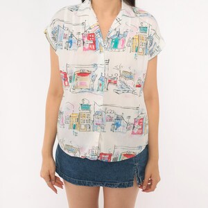 May include: A short-sleeved button-down shirt in white featuring a colorful, hand-drawn style print of various buildings and city structures in shades of red, blue, yellow, and green, paired with a dark blue denim mini skirt with a side slit.