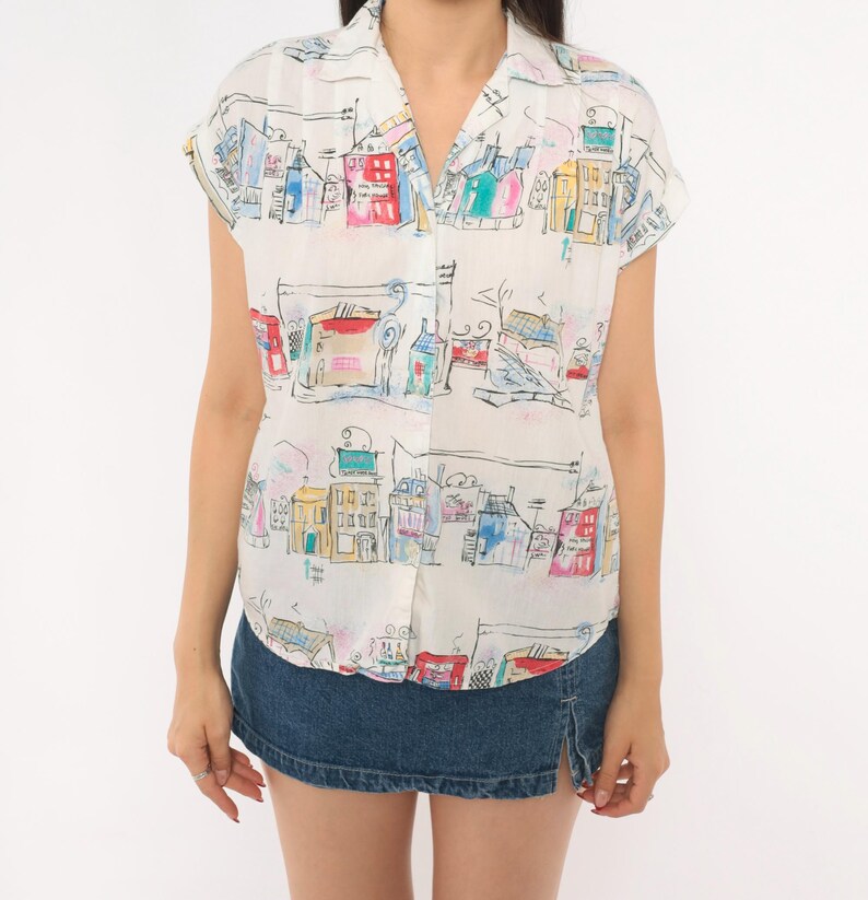 May include: A short-sleeved button-down shirt in white featuring a colorful, hand-drawn style print of various buildings and city structures in shades of red, blue, yellow, and green, paired with a dark blue denim mini skirt with a side slit.