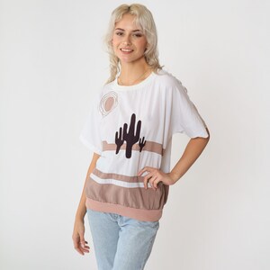 May include: A white t-shirt with a brown and black cactus graphic. The shirt has a relaxed fit and a round neckline.