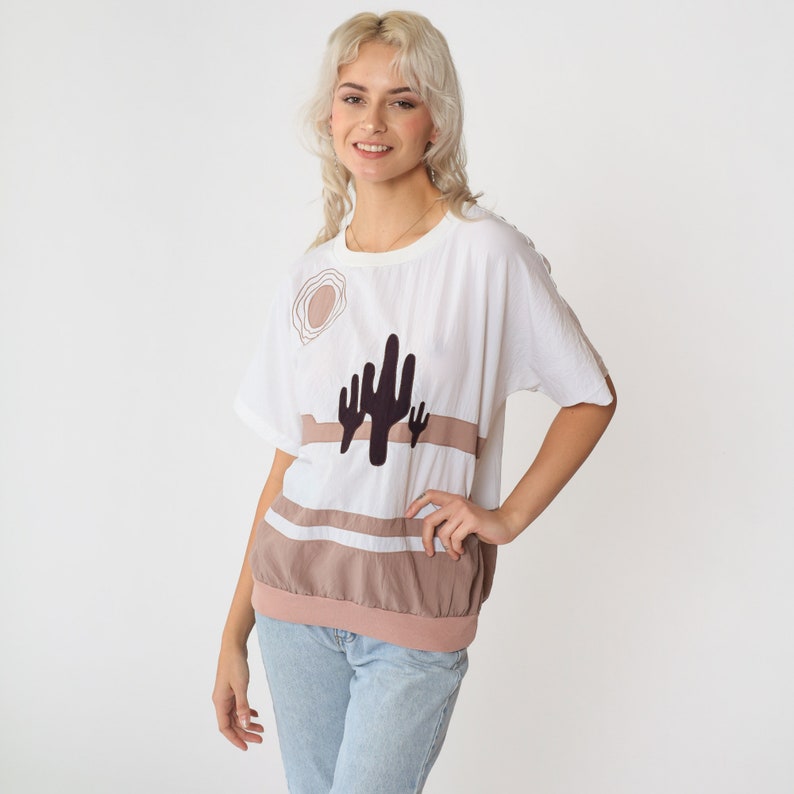 May include: A white t-shirt with a brown and black cactus graphic. The shirt has a relaxed fit and a round neckline.