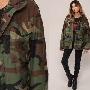 May include: A camouflage field jacket featuring a woodland pattern in shades of green, brown, and black. The jacket includes a front zipper closure, a fold-down collar, and a circular patch on the chest pocket. It is worn over a black shirt.