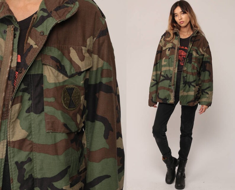 May include: A camouflage field jacket featuring a woodland pattern in shades of green, brown, and black. The jacket includes a front zipper closure, a fold-down collar, and a circular patch on the chest pocket. It is worn over a black shirt.