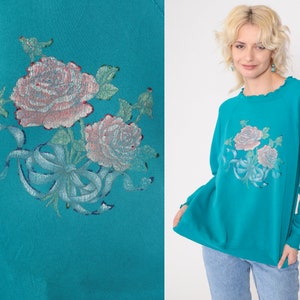 May include: Teal blue sweatshirt with a floral design featuring pink roses and a blue ribbon. The roses are embroidered on the front of the sweatshirt.