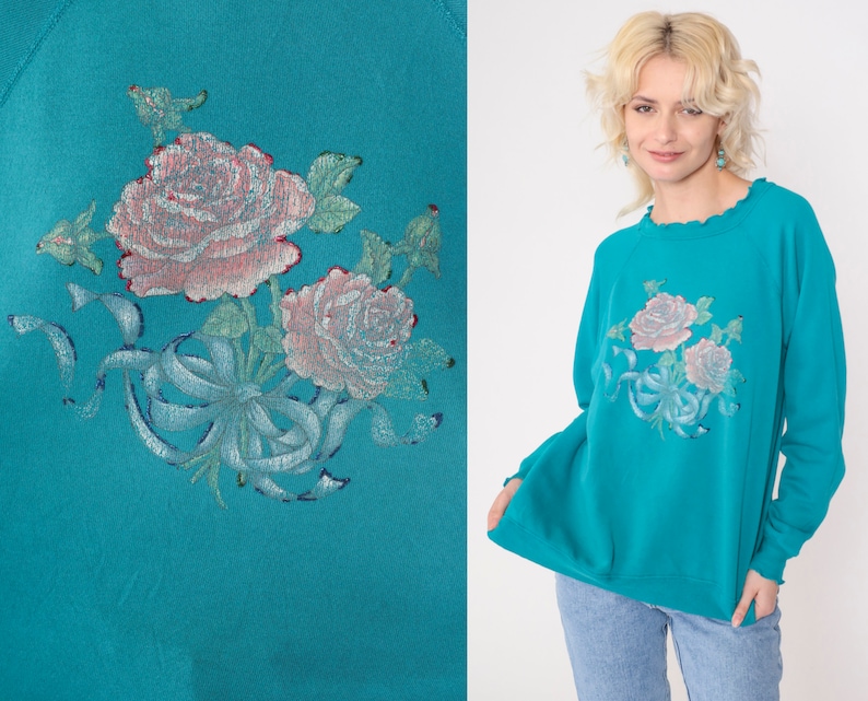 May include: Teal blue sweatshirt with a floral design featuring pink roses and a blue ribbon. The roses are embroidered on the front of the sweatshirt.