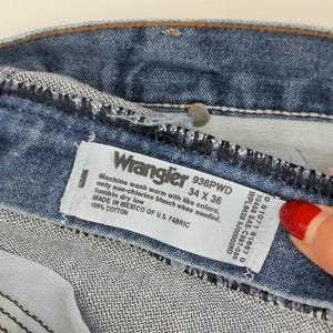 May include: Close-up of blue denim jeans with a Wrangler label. The label indicates a size of 34 x 36 inches. The jeans are made of 100% cotton and manufactured in Mexico of U.S. fabric.