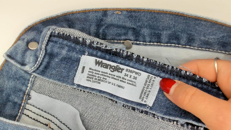 May include: Close-up of blue denim jeans with a Wrangler label. The label indicates a size of 34 x 36 inches. The jeans are made of 100% cotton and manufactured in Mexico of U.S. fabric.