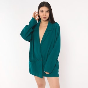 May include: An oversized teal blazer featuring a notched lapel, long sleeves, and two front patch pockets. The garment has a double-breasted design with two visible buttons on the front, providing a structured yet relaxed fit with a deep V-neckline.