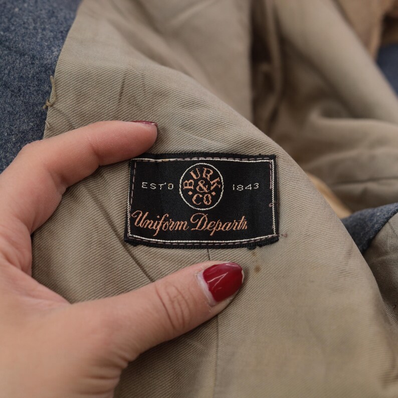 K&ouml;nnte beinhalten: Nahaufnahme des Innenfutters einer Jacke mit einem schwarzen Etikett mit der Aufschrift "Uniform Dept." und dem Firmenlogo. Das Etikett ist auf einem beigefarbenen Futter befestigt, mit einem blauen Au&szlig;enmaterial. Das Etikett lautet "EST'D 1843".