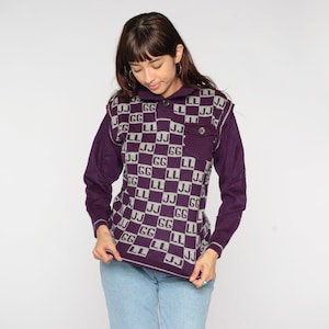 Könnte beinhalten: Eine lila Strickweste mit einem karierten Muster aus grauen "GG" und "LL" Buchstaben. Die Weste hat einen Knopfkrag und eine aufgesetzte Tasche auf der linken Seite.