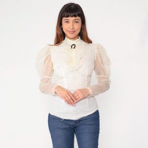 以下が含まれることがあります： クリーム色のブラウス。透け感のあるパフスリーブとハイネックが特徴。ブラウスはフリル付きで、首元には黒い蹄鉄型のディテール。モデルは青いジーンズを着用しています。