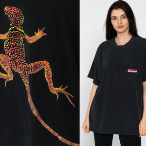 Könnte beinhalten: Schwarzes T-Shirt mit einem farbenfrohen Eidechsen-Design in Gelb, Orange und Pink. Das Shirt hat eine Tasche mit dem Text "MARLBORO UNLIMITED" in Rot und Weiß.