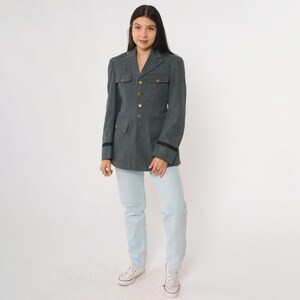 K&ouml;nnte beinhalten: Dunkelgraue Jacke im Milit&auml;rstil mit goldenen Kn&ouml;pfen, schwarzen &Auml;rmelbes&auml;tzen und zwei Vordertaschen. Die Jacke wird mit hellblauen Jeans und wei&szlig;en Turnschuhen getragen. Die Jacke hat eine taillierte Passform.