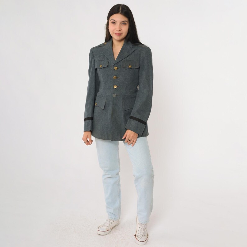 K&ouml;nnte beinhalten: Dunkelgraue Jacke im Milit&auml;rstil mit goldenen Kn&ouml;pfen, schwarzen &Auml;rmelbes&auml;tzen und zwei Vordertaschen. Die Jacke wird mit hellblauen Jeans und wei&szlig;en Turnschuhen getragen. Die Jacke hat eine taillierte Passform.