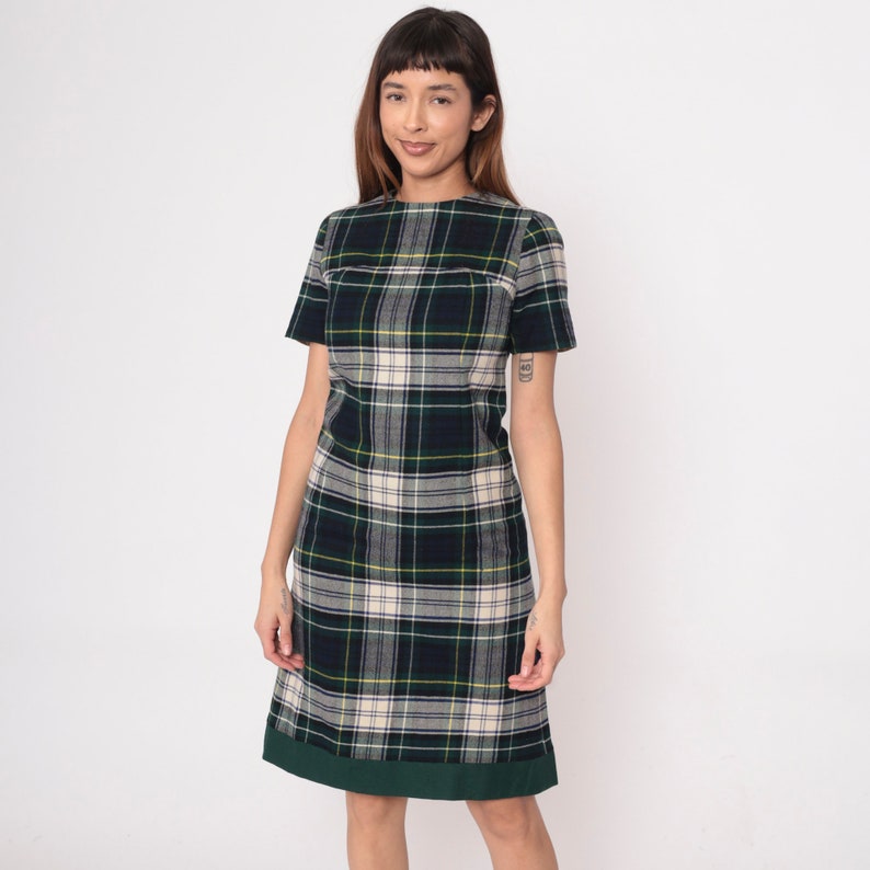 May include: A green, navy, and cream plaid dress with short sleeves. The dress has a straight silhouette and a dark green hemline. The model is wearing the dress in front of a white background.