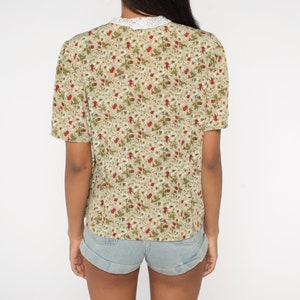 K&ouml;nnte beinhalten: Kurz&auml;rmlige Bluse mit floralem Muster in Gr&uuml;n, Rot und Wei&szlig;. Die Bluse hat einen wei&szlig;en Spitzenkragen und wird mit Jeansshorts kombiniert. Die R&uuml;ckansicht der Bluse wird gezeigt.