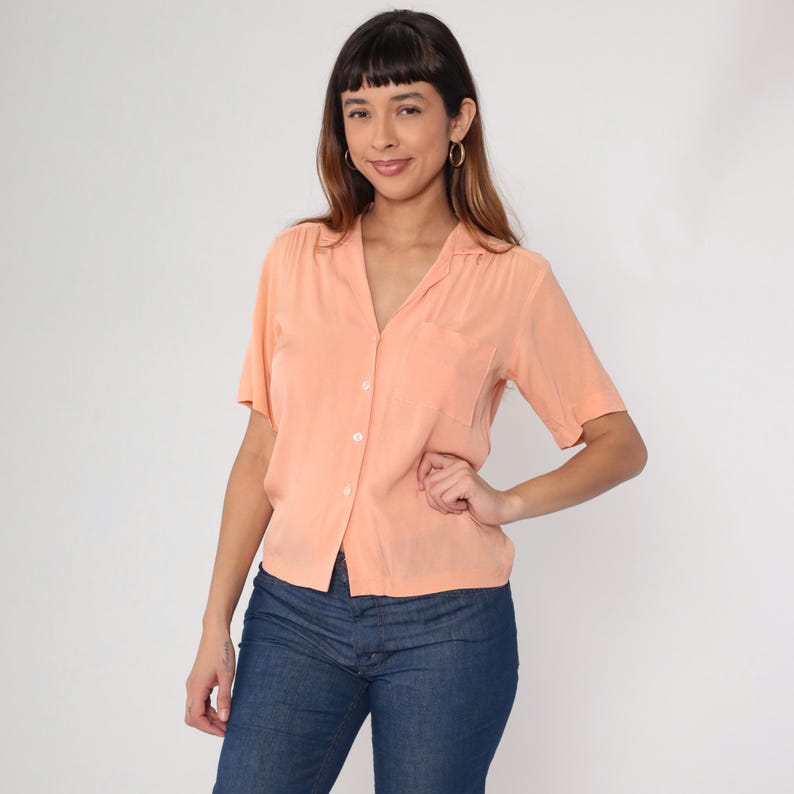 Puede incluir: Una camisa abotonada de color melocot&oacute;n de manga corta con un bolsillo en el pecho izquierdo. La camisa tiene cuello en V y se combina con vaqueros azules. La modelo lleva pendientes de aro dorados.