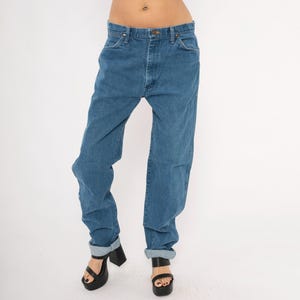 May include: Blue denim jeans with a button and zipper closure. The jeans have a classic five-pocket design and are cuffed at the ankles. The model is wearing black platform sandals. The jeans are a medium wash and appear to be vintage.