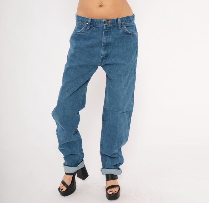 May include: Blue denim jeans with a button and zipper closure. The jeans have a classic five-pocket design and are cuffed at the ankles. The model is wearing black platform sandals. The jeans are a medium wash and appear to be vintage.