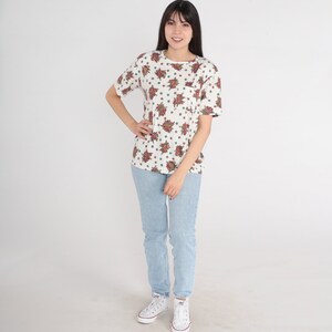 May include: A white t-shirt with a floral print and a light blue pair of jeans. The person is wearing white sneakers.