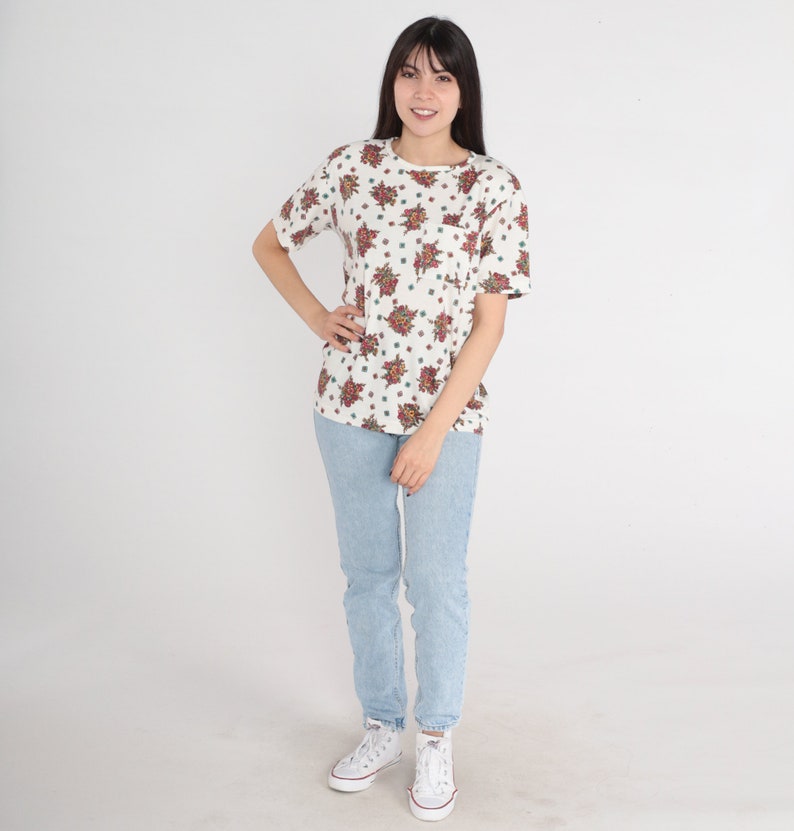 May include: A white t-shirt with a floral print and a light blue pair of jeans. The person is wearing white sneakers.