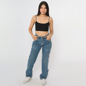 Puede incluir: Una mujer con un top corto negro de tirantes finos y vaqueros azules. Los vaqueros tienen bragueta de botones y un corte relajado. Zapatillas blancas completan el conjunto, sobre un fondo blanco.