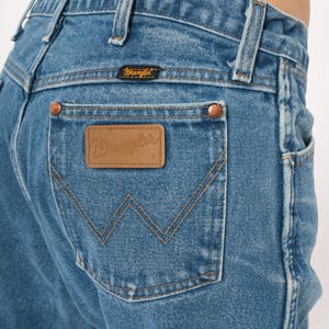 Puede incluir: Primer plano de unos vaqueros de mezclilla azules con un parche de cuero marr&oacute;n con la marca "Wrangler" en el bolsillo trasero. Los vaqueros tienen remaches de cobre y un dise&ntilde;o cl&aacute;sico de cinco bolsillos. La costura es de color beige contrastante.