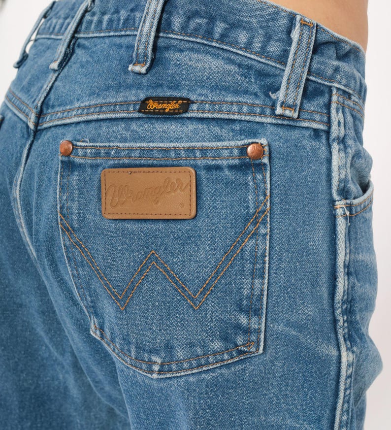 Puede incluir: Primer plano de unos vaqueros de mezclilla azules con un parche de cuero marr&oacute;n con la marca "Wrangler" en el bolsillo trasero. Los vaqueros tienen remaches de cobre y un dise&ntilde;o cl&aacute;sico de cinco bolsillos. La costura es de color beige contrastante.