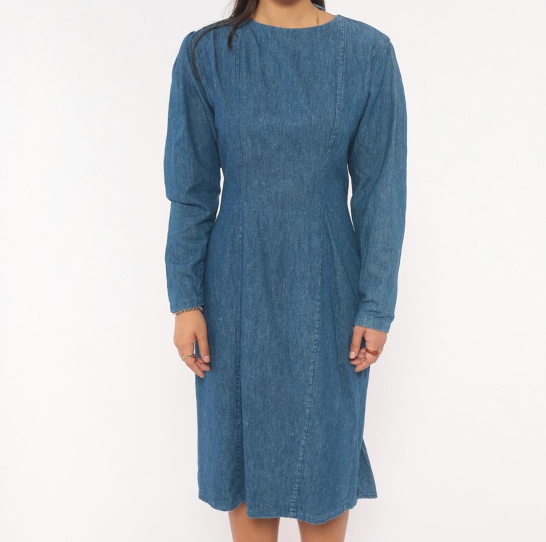 Puede incluir: Un vestido de mezclilla de manga larga en un tono azul medio, con cuello redondo alto, corpi&ntilde;o ajustado con costuras verticales en la parte delantera y una falda de largo midi con una silueta recta y estructurada.