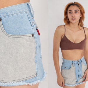 Puede incluir: Pantalones cortos de mezclilla azul claro con un panel gris claro en la parte delantera. Los pantalones cortos tienen un dobladillo deshilachado y una cintura alta.