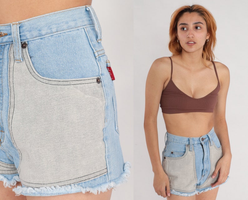 Puede incluir: Pantalones cortos de mezclilla azul claro con un panel gris claro en la parte delantera. Los pantalones cortos tienen un dobladillo deshilachado y una cintura alta.