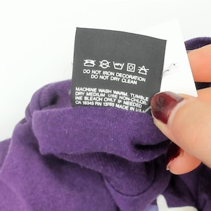 May include: A close-up of a purple garment with a care label. The black label displays washing instructions, including "DO NOT IRON DECORATION" and "DO NOT DRY CLEAN." The label also includes information about machine washing and tumble drying.