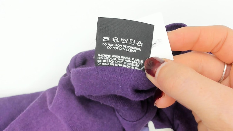 May include: A close-up of a purple garment with a care label. The black label displays washing instructions, including "DO NOT IRON DECORATION" and "DO NOT DRY CLEAN." The label also includes information about machine washing and tumble drying.