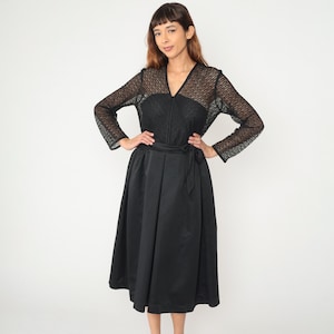 Puede incluir: Un vestido negro con escote en V y mangas largas. El corpiño presenta un diseño de encaje, y la falda es de tela lisa y fluida. Un cinturón a juego ciñe la cintura. El vestido es hasta la rodilla.