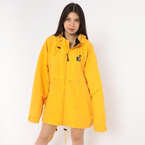 May include: A bright yellow hooded jacket featuring a full-length front zipper, a black interior lining, and a small black square patch with the Carhartt logo on the left chest. The jacket includes adjustable drawstrings at the hood and hem.