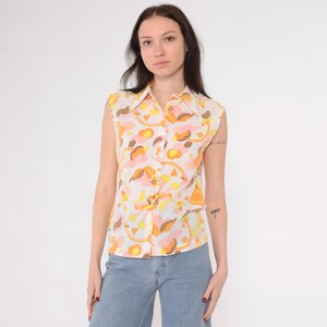 May include: A sleeveless button-down shirt with a colorful abstract pattern in shades of orange, pink, yellow, and brown. The shirt has a collared neckline and is made of a lightweight fabric. The shirt is paired with light blue denim jeans.