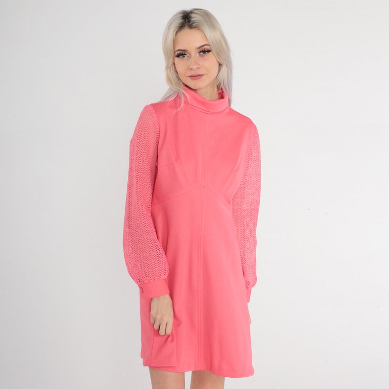 May include: A pink dress with a high neck and long sleeves. The sleeves are made of a sheer, lacy material. The dress is fitted through the bodice and flares out slightly at the skirt.