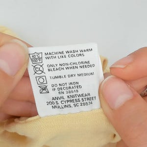 May include: A close-up of a clothing care label held between two hands. The label is white with black text and symbols, including washing instructions, temperature, and the brand name "ANVIL KNITWEAR".