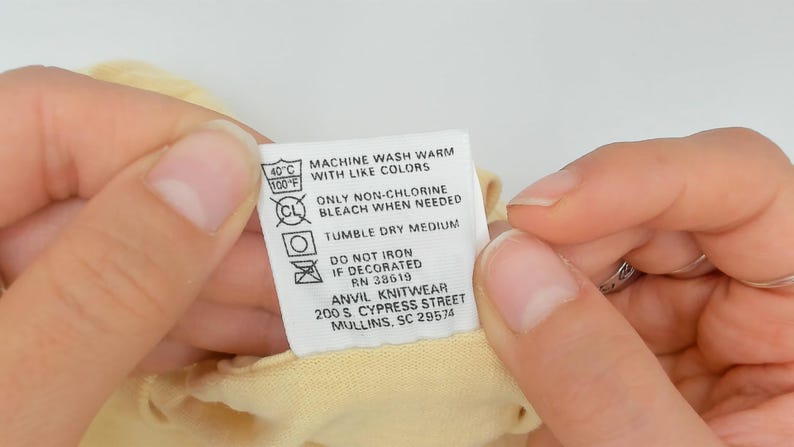 May include: A close-up of a clothing care label held between two hands. The label is white with black text and symbols, including washing instructions, temperature, and the brand name "ANVIL KNITWEAR".