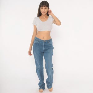 May include: A light gray crop top and blue jeans. The crop top has short sleeves and a scoop neck. The jeans are a medium wash denim with a straight leg. The model is standing in front of a white background.