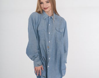 May include: A light blue long-sleeve shirt with two chest pockets and a button-down front. The shirt has a classic collar and is worn with blue jeans. The shirt has paint splatters on the front and sleeves.
