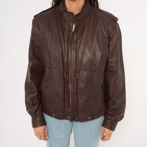 May include: A dark brown leather jacket with a zip-up front, a stand-up collar, and shoulder epaulets. The jacket has long sleeves and a slightly textured appearance. The bottom hem has two snap closures. The model is wearing light blue jeans.