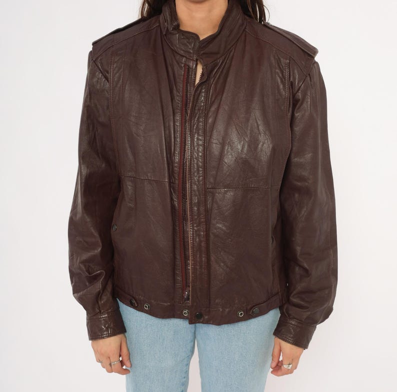 May include: A dark brown leather jacket with a zip-up front, a stand-up collar, and shoulder epaulets. The jacket has long sleeves and a slightly textured appearance. The bottom hem has two snap closures. The model is wearing light blue jeans.