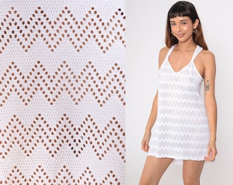 May include: White crochet dress with a V-neck and crisscross straps. The dress features a chevron pattern with openwork details. The model is wearing the dress. The dress is a mini length.