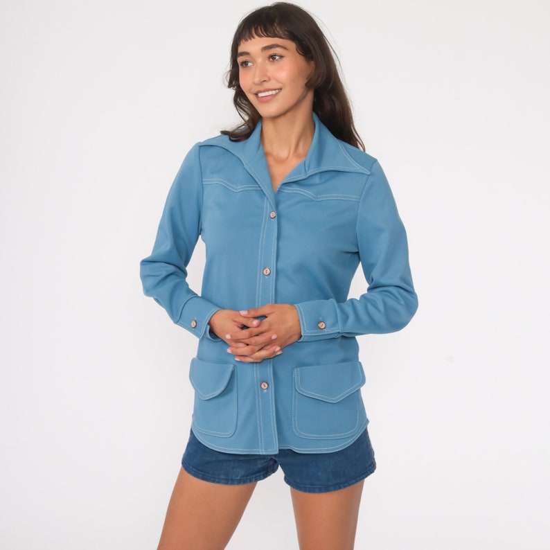 May include: A light blue long-sleeved shirt with a collared neckline, button closure, and two patch pockets. The shirt has white stitching details.