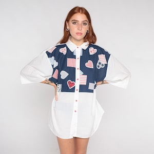 May include: A white button-down shirt with a blue panel featuring a patchwork design of red and white hearts and squares.